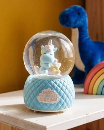 Blue unicorn snow globe on a quilted light-blue base with 'Enjoy and Dream' label, pastel rainbow toys blurred in the background.