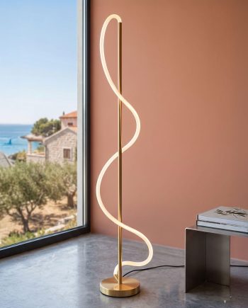 Gold spiral LED floor lamp with warm light standing on a circular base beside a window, overlooking a coastal village and blue sea.
