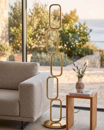 Tall gold geometric sculpture with stacked rounded-rectangle links on a circular base beside a beige sofa by a window.