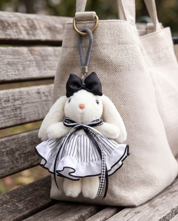 Plush white bunny keychain with a black bow, striped dress, hanging from a beige bag on a wooden bench