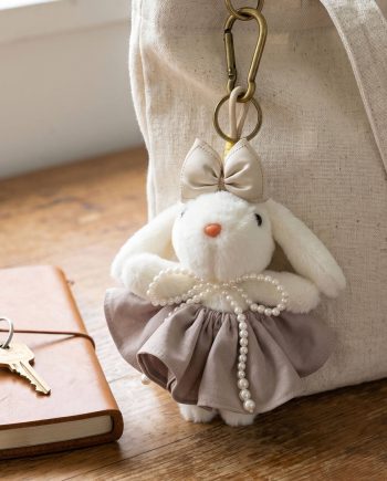 White plush bunny keychain with a beige bow and pearl-bead necklace hanging from a tan bag, on a wooden surface near a notebook and keys.