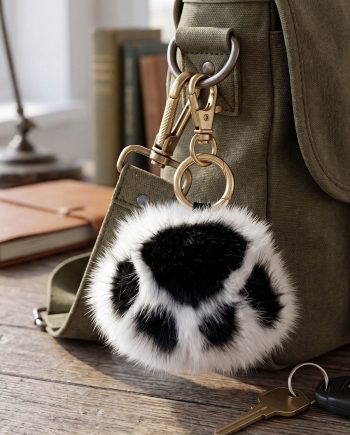 Green canvas backpack with gold hardware and a black-and-white fur pom-pom keychain hanging from a ring, on a wooden surface with blurred books in the background.