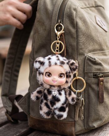 Leopard-print plush keychain hanging from a khaki backpack zipper with gold rings and keys beside a small animal figure