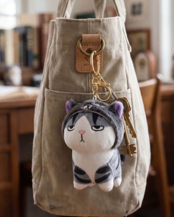 Beige canvas bag with a leather tag and gold key ring holding a gray-and-white plush cat keychain.