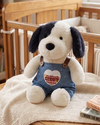 Soft plush dog with dark ears wearing blue denim overalls sits on a cream knit blanket in a cozy nursery. A wooden crib and stacked books are in the background.