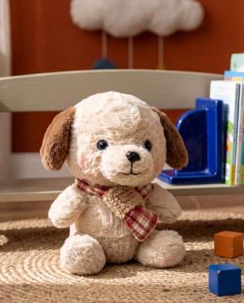 Soft plush puppy with a red plaid scarf sits on a woven rug in a child’s play area, with books and blocks nearby.