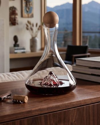 Glass wine decanter with a wooden stopper on a wooden table, a corkscrew nearby, and mountains visible through the window in the background.