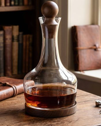 Glass decanter with a wooden stopper on a wooden table, amber liquid inside, a library backdrop in soft focus.