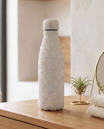 Tall silver rhinestone-covered bottle on a wooden dresser, with a small potted plant and towel in the background.