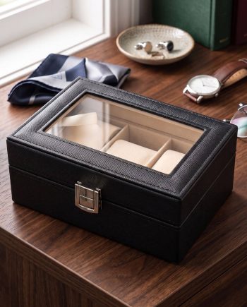 Black leather jewelry box with a glass top on a wooden table, open to beige cushioned compartments.